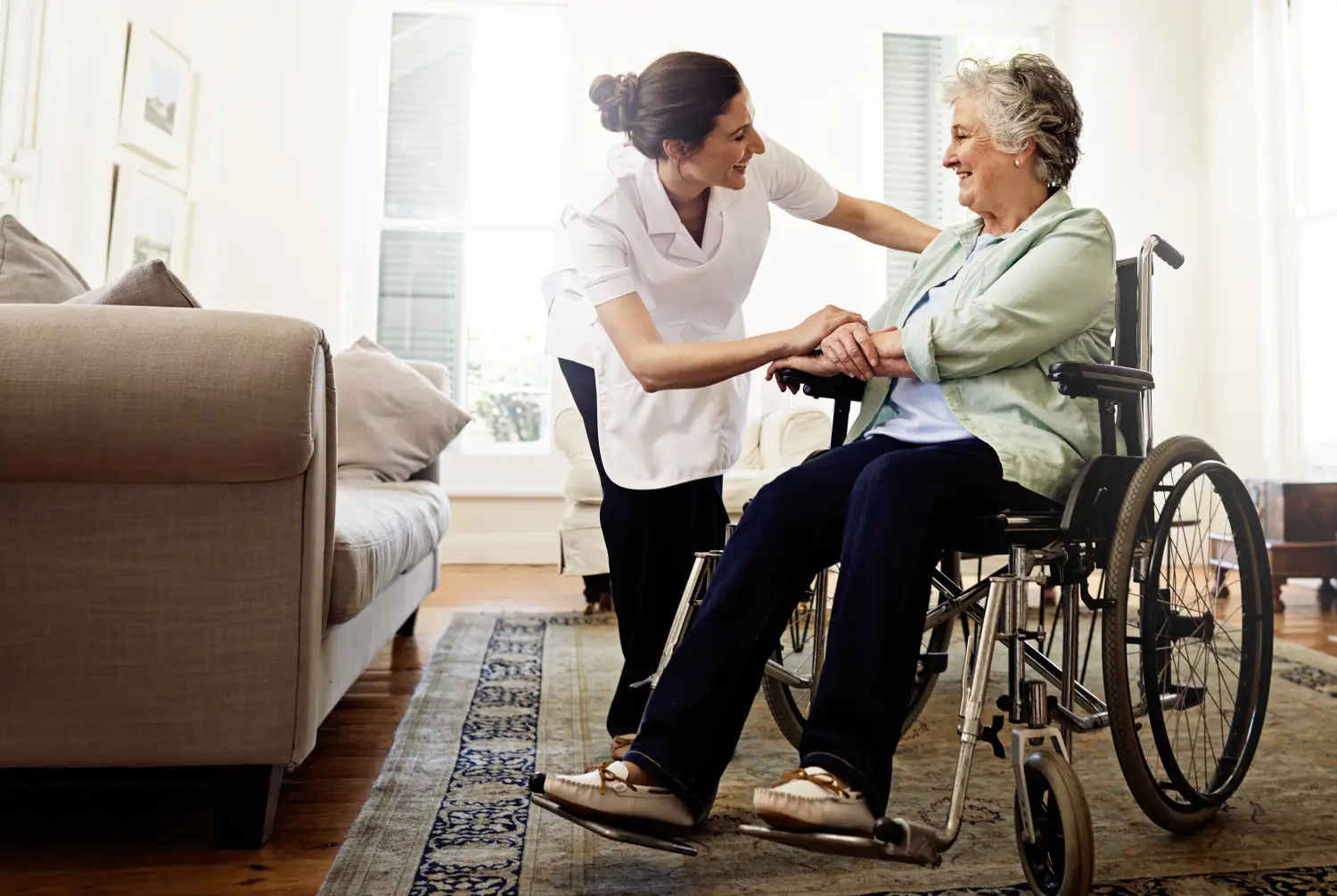 Aufnahme einer lächelnden Pflegekraft, die einer älteren Frau im Rollstuhl zu Hause hilft.